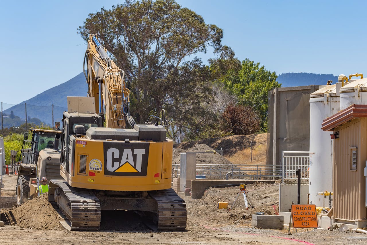 contact-img Excavator at a construction site with a road closed sign and mountainous backdrop.