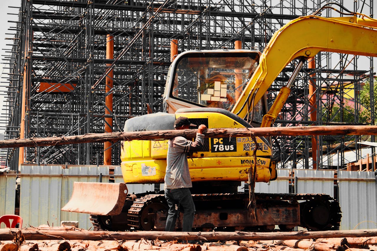 gallery-6 A construction worker manages logs with an excavator at a busy industrial site with scaffolding.