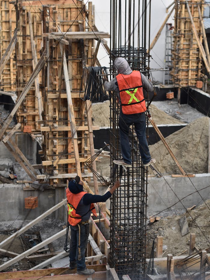 gallery-4 Two workers in safety vests climb a steel structure on a busy construction site.