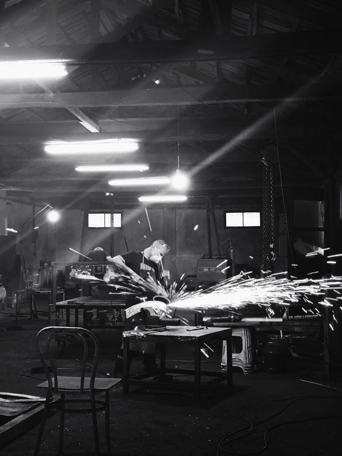 gallery-1 A black and white photo of a workshop with a worker welding, emitting bright sparks.